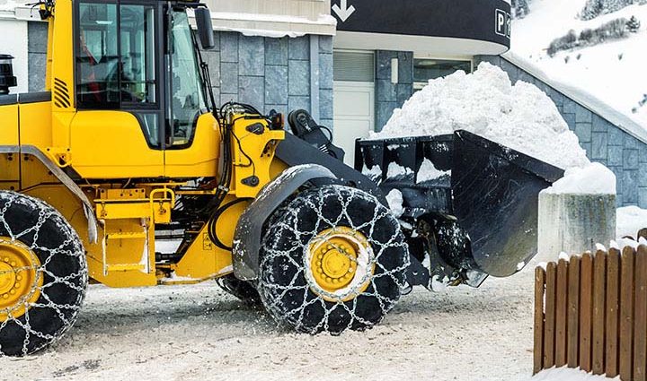 Plowing Through Winter Weather: Snow Removal You Can Rely On; Big loader machine with steel metal chains removing big snow pile from city street at alpine mountain region in winter. Heavy snowfall aftermath. precipitation cleaning equipment.