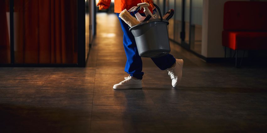 Janitorial staff carrying cleaning bucket through dim hallway