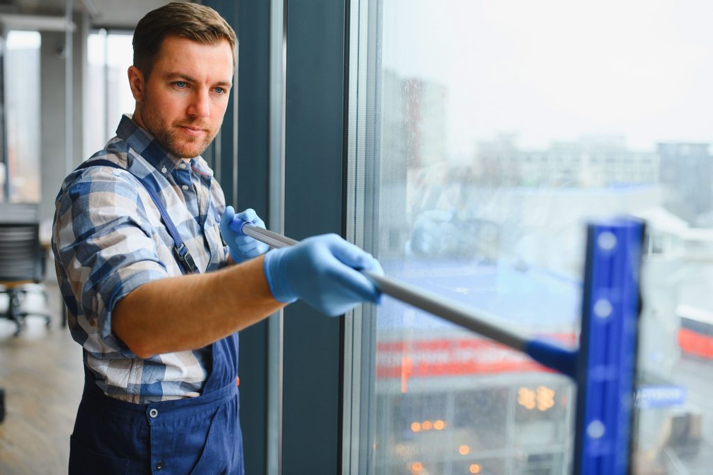 Commercial cleaner squeegeeing large office window with blue equipment